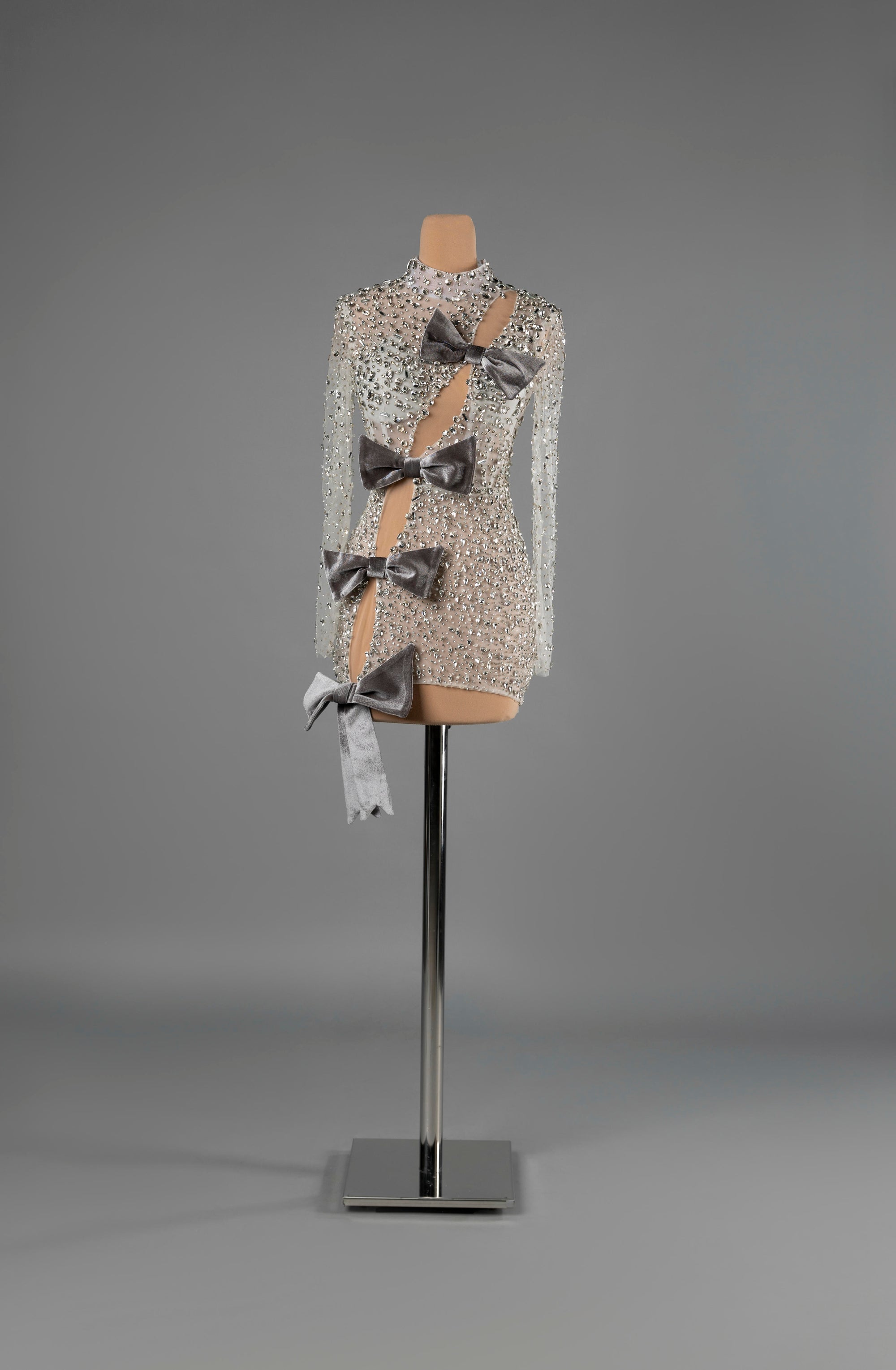 Sihëna Argent Bows — silver high-neck long-sleeve fitted mini dress, crystal-beaded over sheer nude with a diagonal cutout and grey velvet bows.