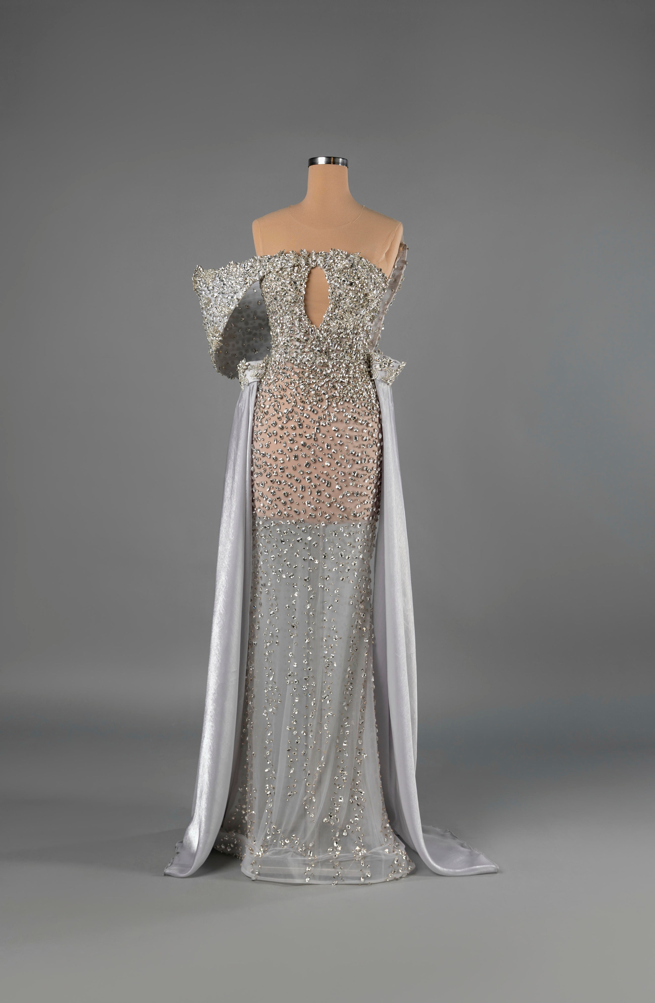 Sihëna Ivory Regalia — silver strapless mermaid gown densely crystal-beaded over sheer nude with structured off-shoulder sleeves and chiffon cape panels.