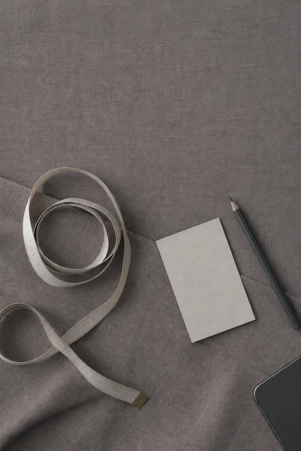A soft cream tape with faint markings, loosely coiled on mauve-grey linen, beside a blank card, a pencil and a black phone.
